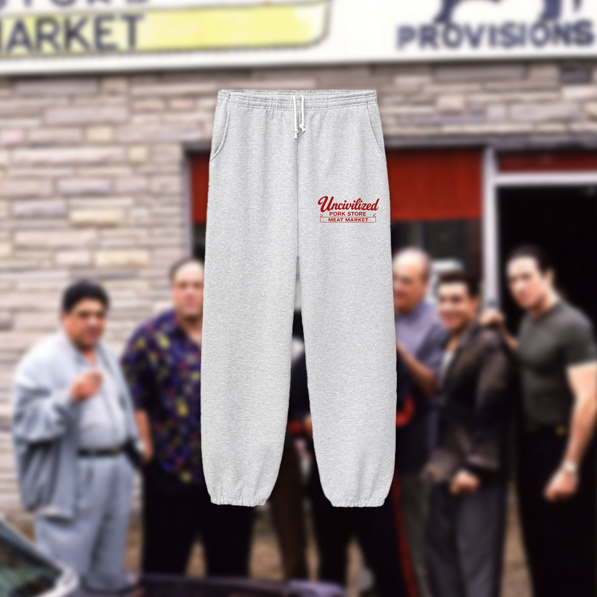 Light gray sweatpants with a red screen printed logo reading 'Uncivilized' and smaller text below, shown floating against a blurred outdoor storefront background with people.